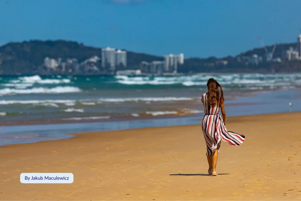 Woman walking along the golden shoreline of Miami Beach, Gold Coast, Queensland, with ocean waves and Burleigh Headland in the background.