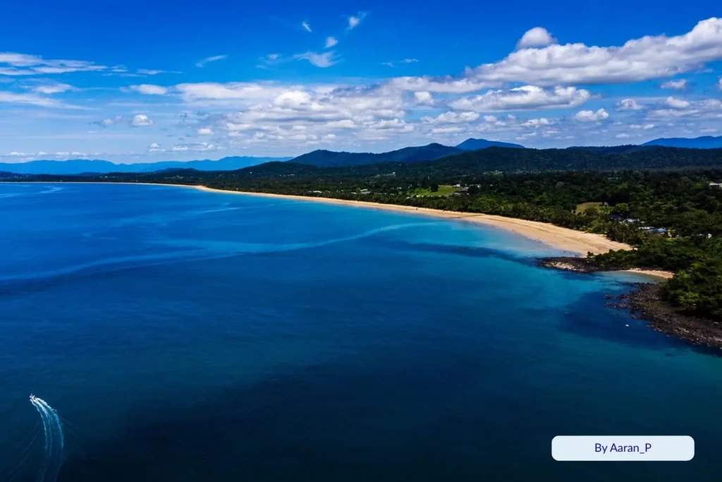 Aerial panorama of Mission Beach showing golden coastline, rainforest-covered hills, and deep blue Coral Sea, Cassowary Coast, Queensland, Australia.