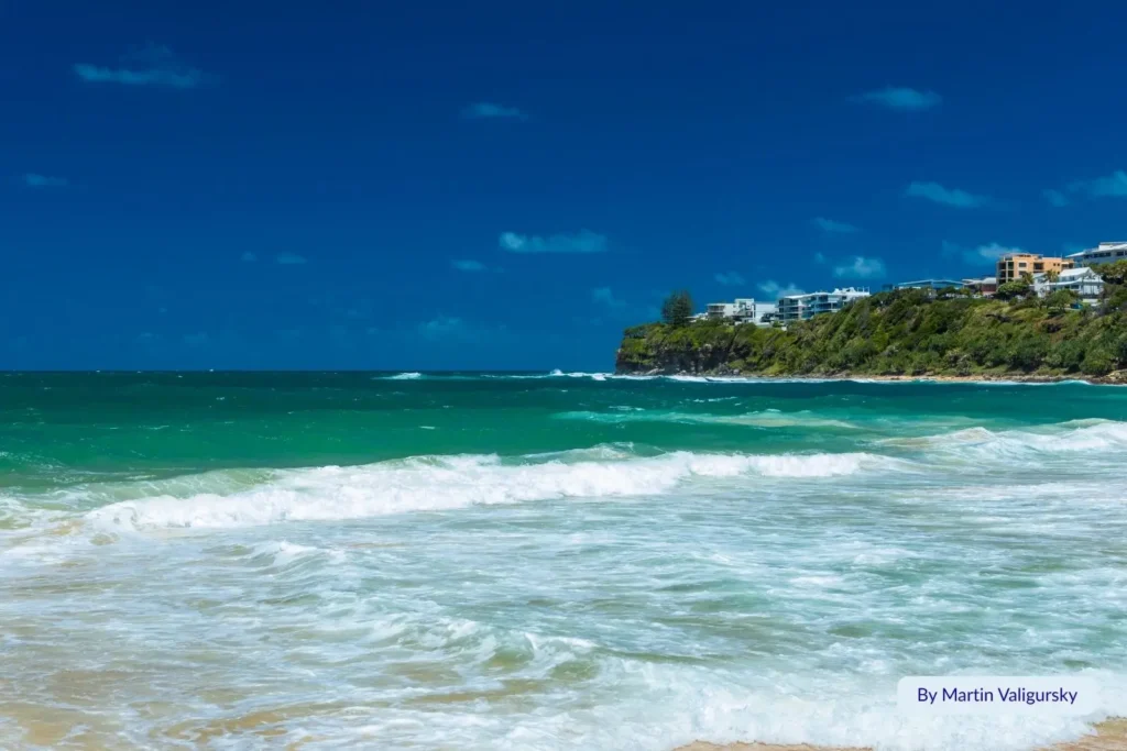 Turquoise waves and coastal headland at Moffat Beach, Sunshine Coast, Queensland — surf rolling toward shore with clifftop apartments and blue summer sky in the background.