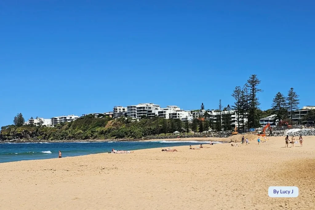 Relaxed day at Moffat Beach, Sunshine Coast, Queensland, with swimmers and sunbathers on the golden sand and coastal apartments in the background.