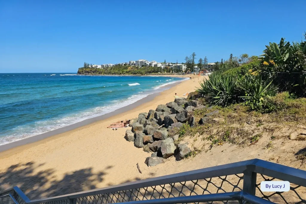 Scenic view of Moffat Beach, Caloundra, Sunshine Coast, Queensland, with golden sand, coastal vegetation, rocky foreshore, and ocean waves under clear blue skies.