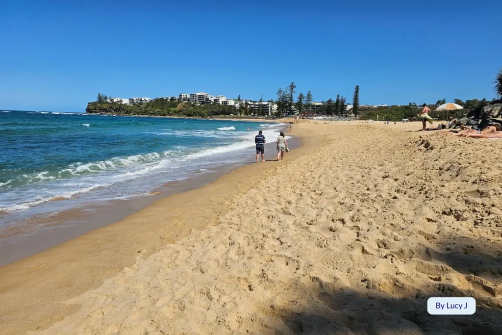 Scenic shoreline of Moffat Beach, Sunshine Coast, Queensland, with golden sand and people walking along the gentle surf under a clear blue sky.