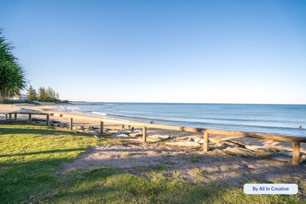 Scenic foreshore view of Moffat Beach, Caloundra, Sunshine Coast, Queensland — calm ocean waves meeting golden sand beside grassy picnic areas and timber fencing.