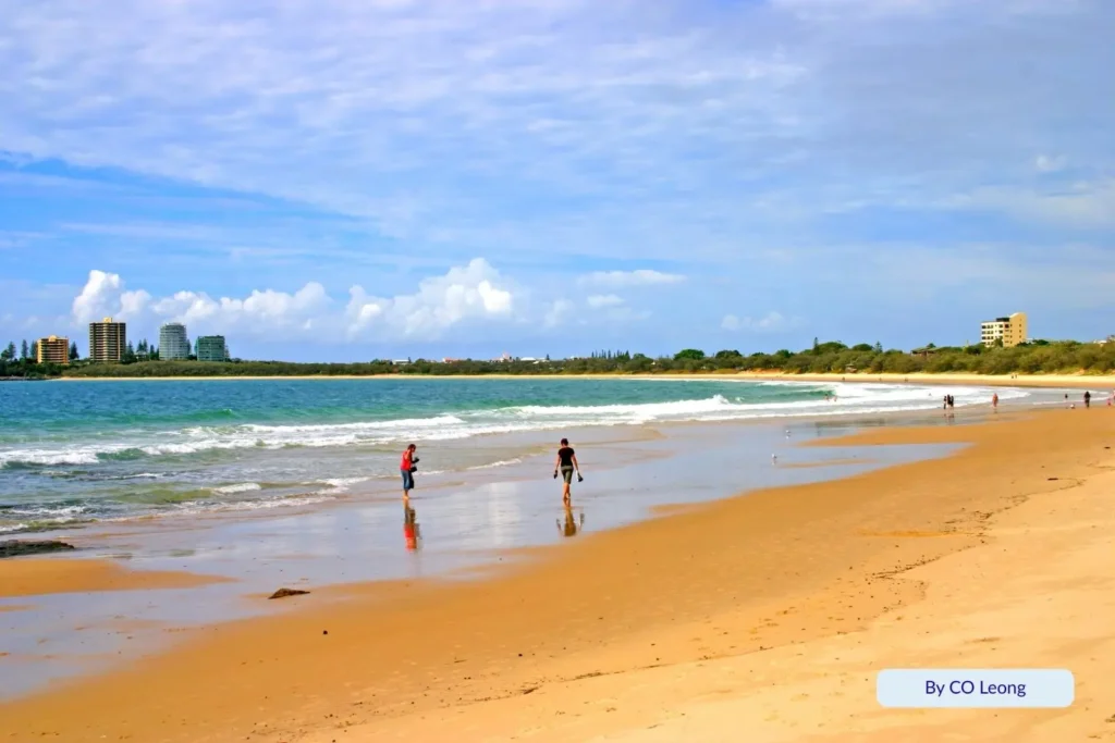 People wading in shallow waves at Mooloolaba Beach, Sunshine Coast, Queensland — golden sand, calm surf, and apartment buildings in the background under a partly cloudy sky.
