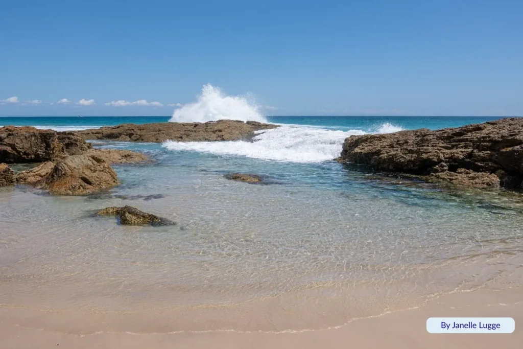 Ocean waves crashing over volcanic rocks at Champagne Pools, Moreton Island, creating natural bubbling pools beneath a clear blue sky.
