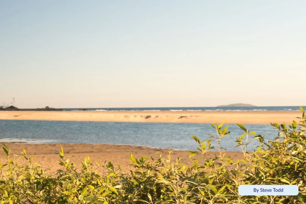 Golden sand and calm tidal waters at Mulambin Beach near Yeppoon, Queensland, viewed across the dune vegetation with Great Keppel Island visible on the horizon.