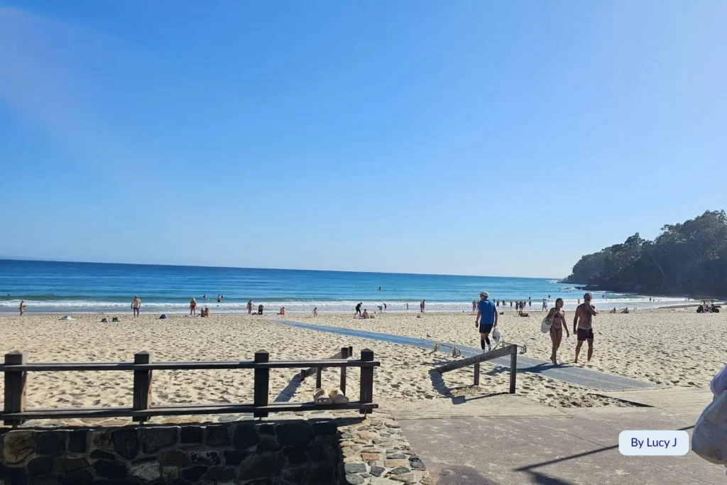 Visitors enjoying a sunny day on the golden sand of Noosa Heads Main Beach, Sunshine Coast, Queensland, with calm waves and forested headland in the distance.