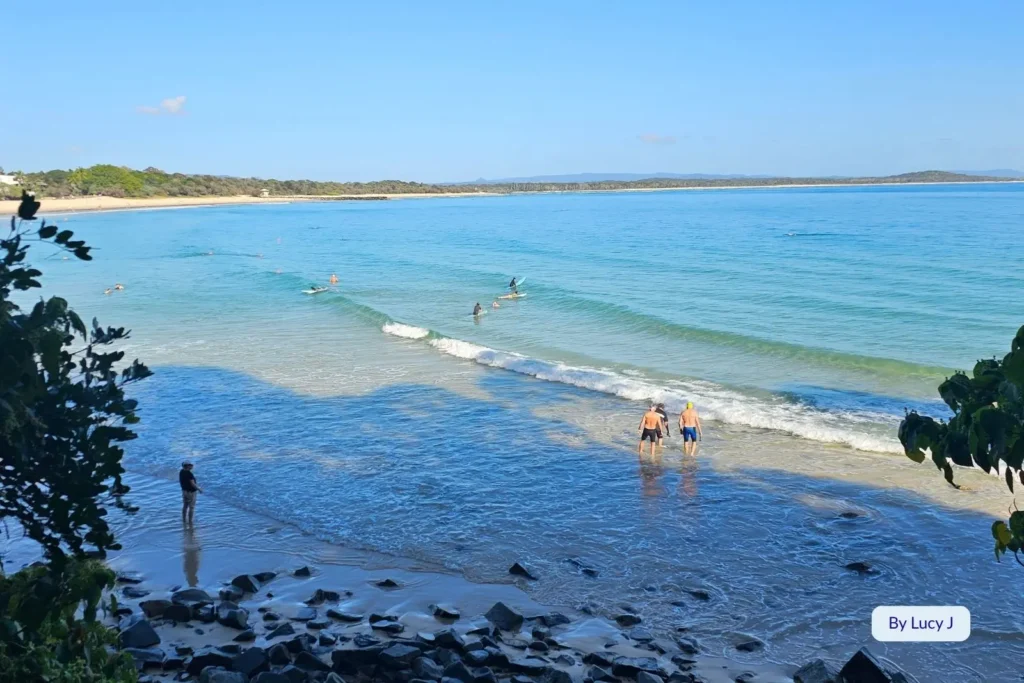 Swimmers and surfers in the gentle turquoise waters at Noosa Heads Main Beach, Sunshine Coast, Queensland, framed by leafy coastal trees and scenic views.