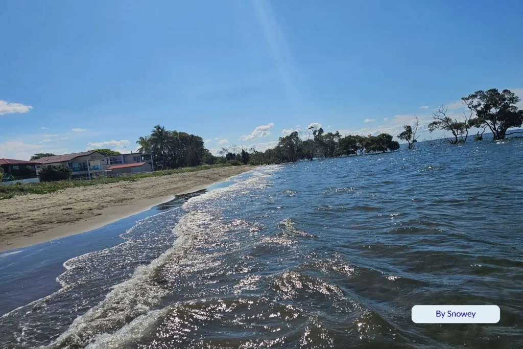 Gentle waves lapping the sandy foreshore at Nudgee Beach with houses and trees along the waterfront, Brisbane.