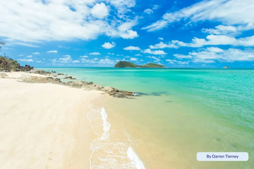 Golden sand and clear turquoise water at Palm Cove, Cairns, Queensland, with rocky headland and Double Island visible in the distance beneath a sunny sky.