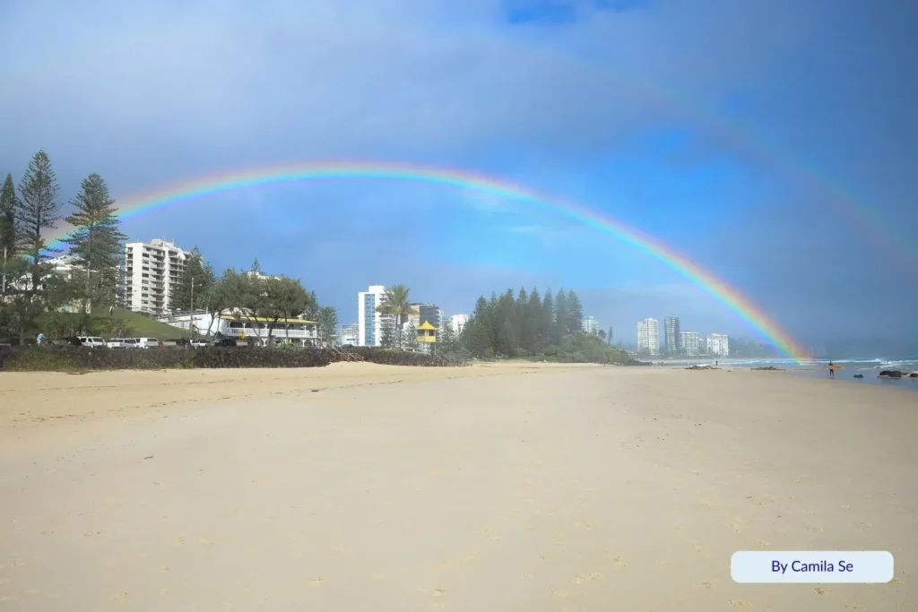 Rainbow arching over Rainbow Bay Beach with golden sand, ocean waves, and beachfront apartments under a bright sky, Gold Coast, Queensland.