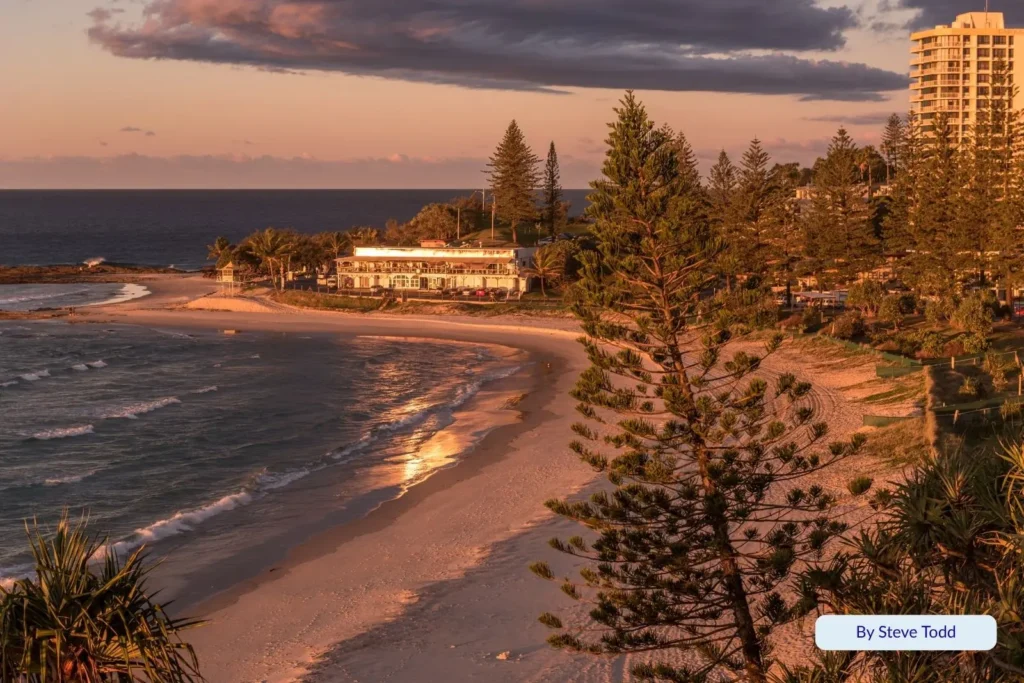 Sunset glow over Rainbow Bay with golden reflections on the sand, pine trees along the foreshore, and the surf club overlooking the beach, Gold Coast, Queensland.