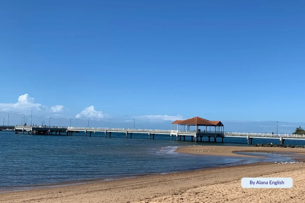 Redcliffe Jetty extending over calm blue waters with golden sandy beach in the foreground under a clear Queensland sky, Moreton Bay, Brisbane