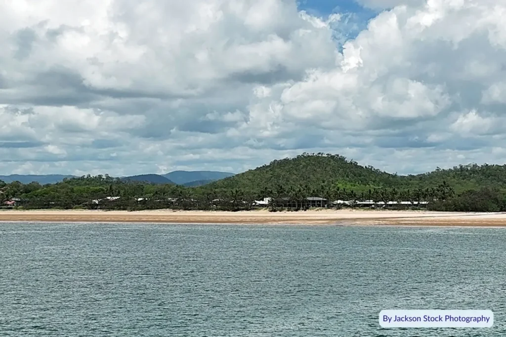 Coastal view of Sarina Beach, Queensland, with lush green headland and calm blue water under a partly cloudy sky.