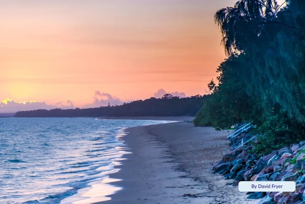 Peaceful sunset at Scarness Beach, Hervey Bay, with gentle waves washing the shoreline and trees silhouetted against a colourful sky.