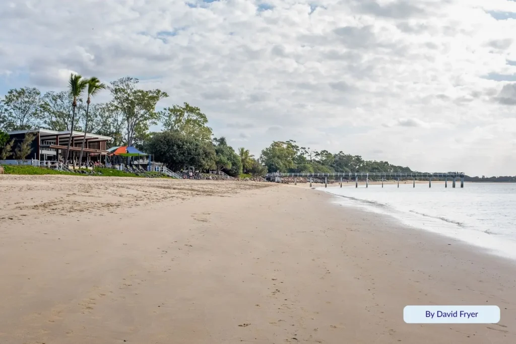 Scarness Beach in Hervey Bay, Queensland, with calm water, sandy foreshore, and waterfront cafés surrounded by palm trees under a cloudy sky.
