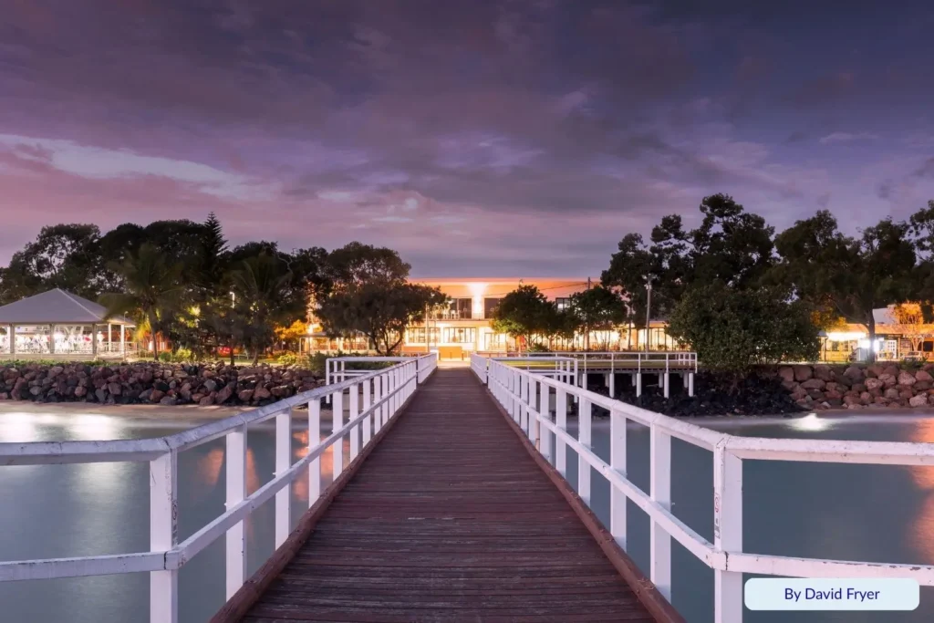 Scarness Jetty in Hervey Bay illuminated at dawn, showing reflections on the water and lights from nearby restaurants and palm-lined foreshore.