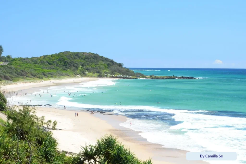 Shelly Beach, Caloundra, Queensland, with turquoise waves and green headland — swimmers enjoying calm surf framed by pandanus palms and lush coastal vegetation.
