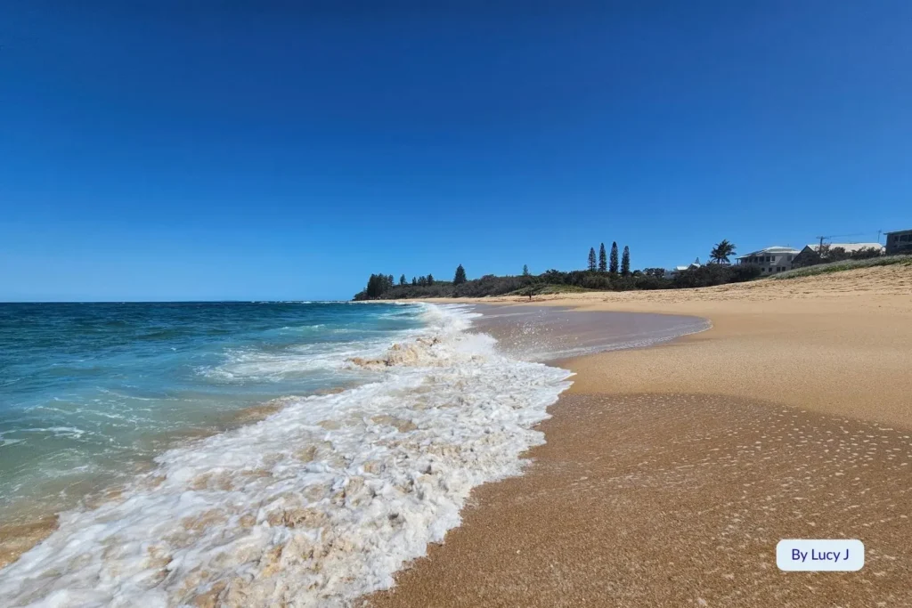 Turquoise surf washing over the sandy foreshore at Shelly Beach, Sunshine Coast, Queensland, with distant trees and coastal homes under bright summer skies.