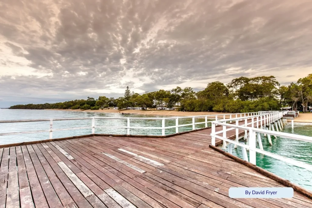 Wooden jetty stretching into the calm turquoise water at Shelley Beach, Torquay, Hervey Bay, Queensland, with trees along the foreshore under a cloudy sky.