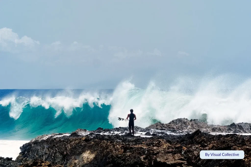 Surfer standing on rocks watching a massive turquoise wave crash at Snapper Rocks, Gold Coast, under bright blue skies.