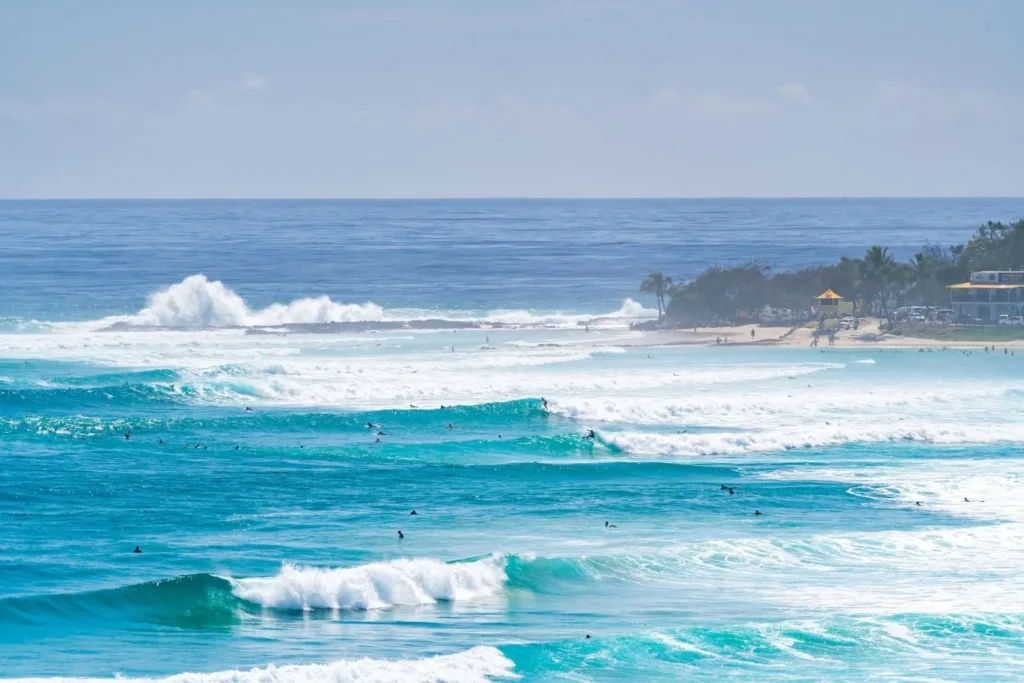 Turquoise surf rolling into Snapper Rocks, with surfers scattered across the lineup and Froggies Beach visible in the distance, Gold Coast, Queensland.