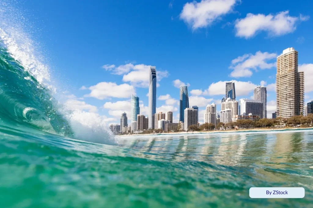 Wave curling near the shore with the Surfers Paradise skyline and high-rise buildings gleaming under a bright Queensland sky.
