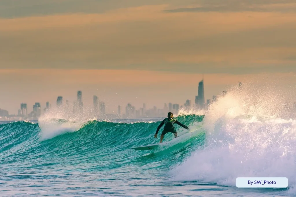 Surfer catching a golden sunrise wave with Surfers Paradise skyscrapers visible in the distance across the shimmering water.