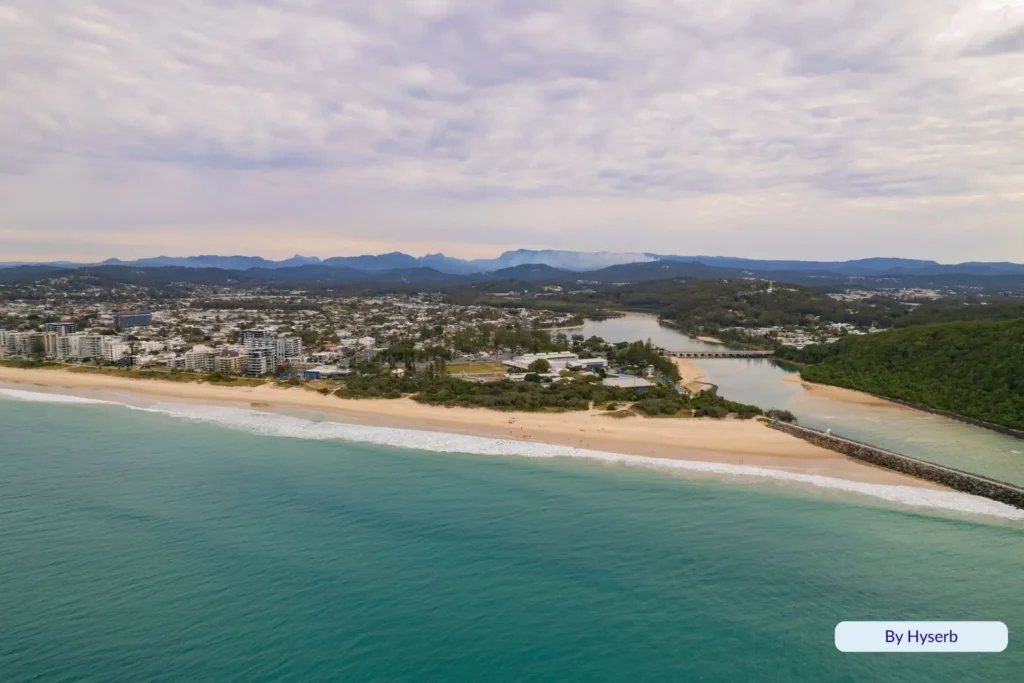 Aerial view of Tallebudgera Beach and creek inlet with golden sand, turquoise surf, lush headland, and the Gold Coast skyline beneath a soft overcast sky.