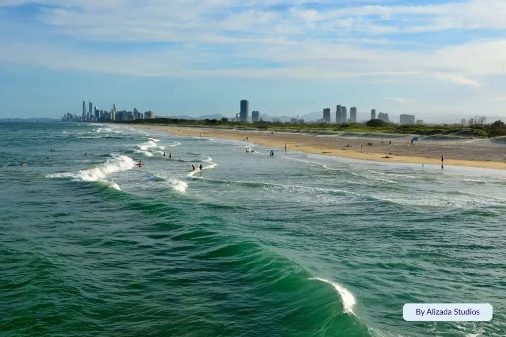 Surfers and swimmers enjoying small waves at The Spit, Gold Coast, with city skyscrapers and beachfront parklands in the background.