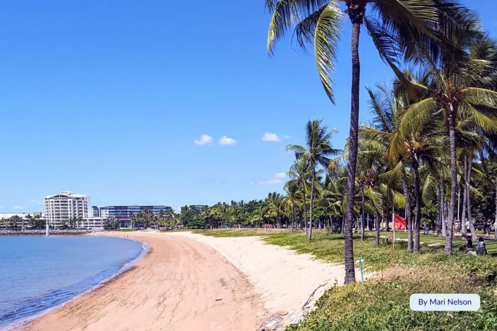 Curved sandy shoreline and palm-lined promenade at The Strand, Townsville, with city buildings in the background, Queensland, Australia.