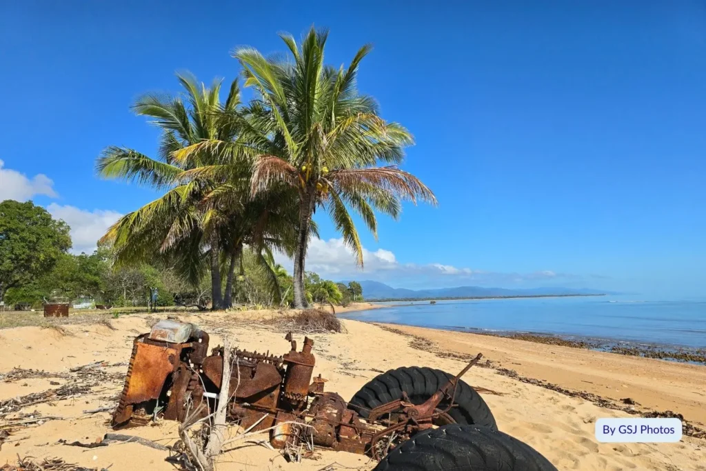 Rusted tractor wreck and palm trees on the sandy shoreline of Toolakea Beach overlooking tranquil coastal waters in North Queensland.
