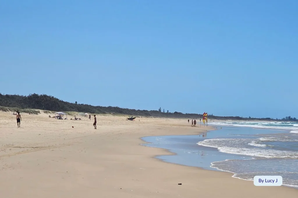 Wide sandy stretch of Wurtulla Beach, Sunshine Coast, Queensland, with gentle surf, beach walkers, and lifeguard patrol under clear blue skies.