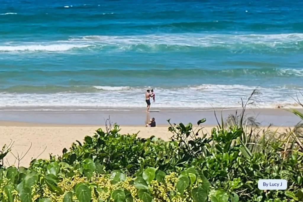 Family enjoying the shoreline at Wurtulla Beach, Sunshine Coast, Queensland, with turquoise waves rolling in beyond lush green coastal dunes.