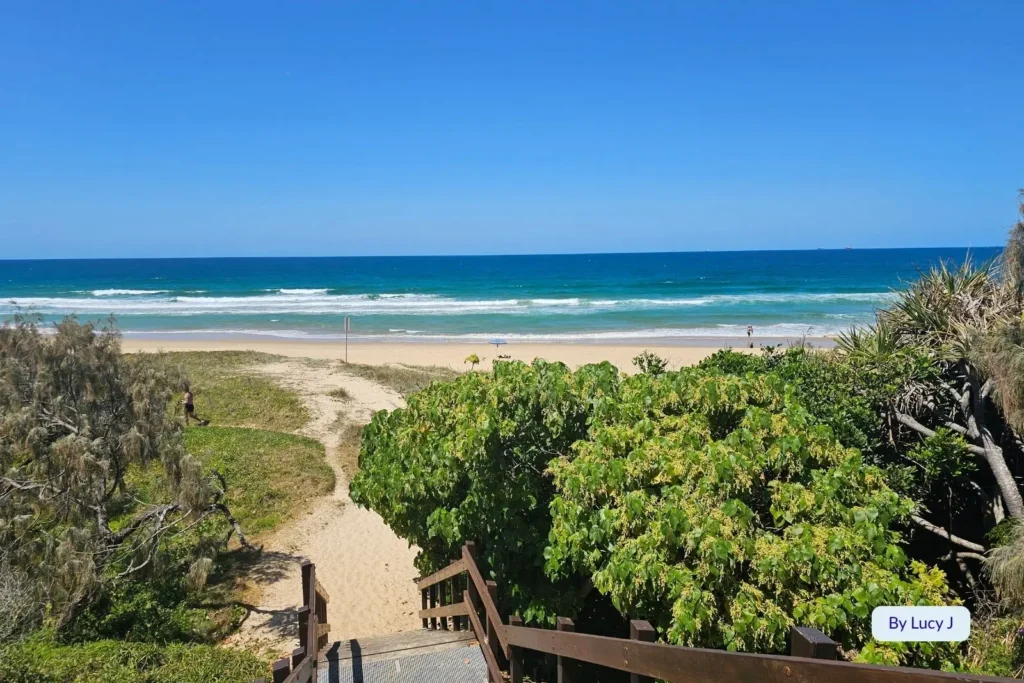 Wooden boardwalk leading through green coastal vegetation to the golden sands and blue surf of Wurtulla Beach, Sunshine Coast, Queensland.