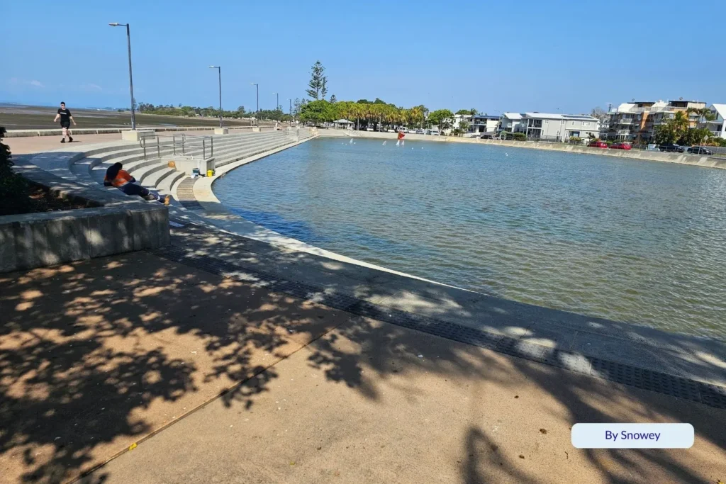 The Wynnum tidal wading pool in Brisbane, Queensland, with calm water, shaded seating areas, and families enjoying the sunny bayside.