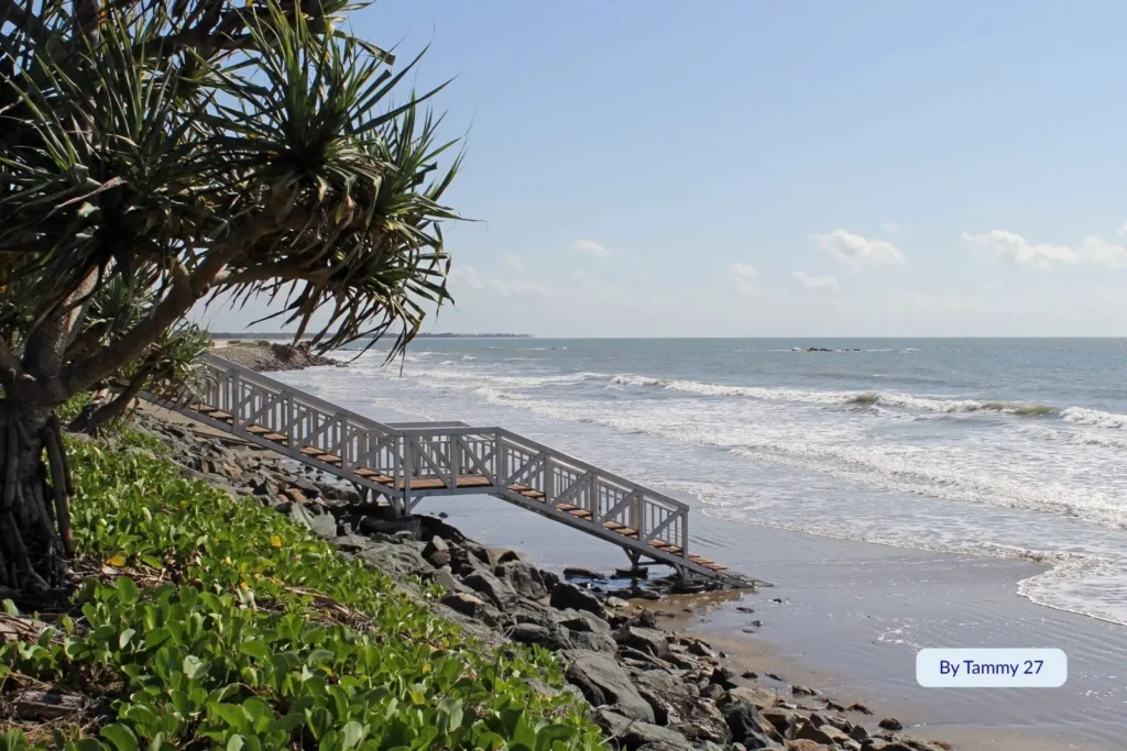 Boardwalk and pandanus trees along Yeppoon Main Beach with waves rolling in under a bright blue sky