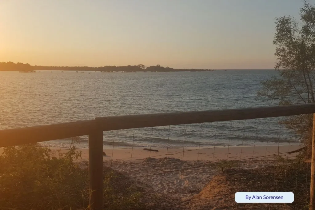 Golden sunset over Toogoom Beach near Hervey Bay, Queensland, viewed through a timber fence with gentle waves lapping the sandy shore.
