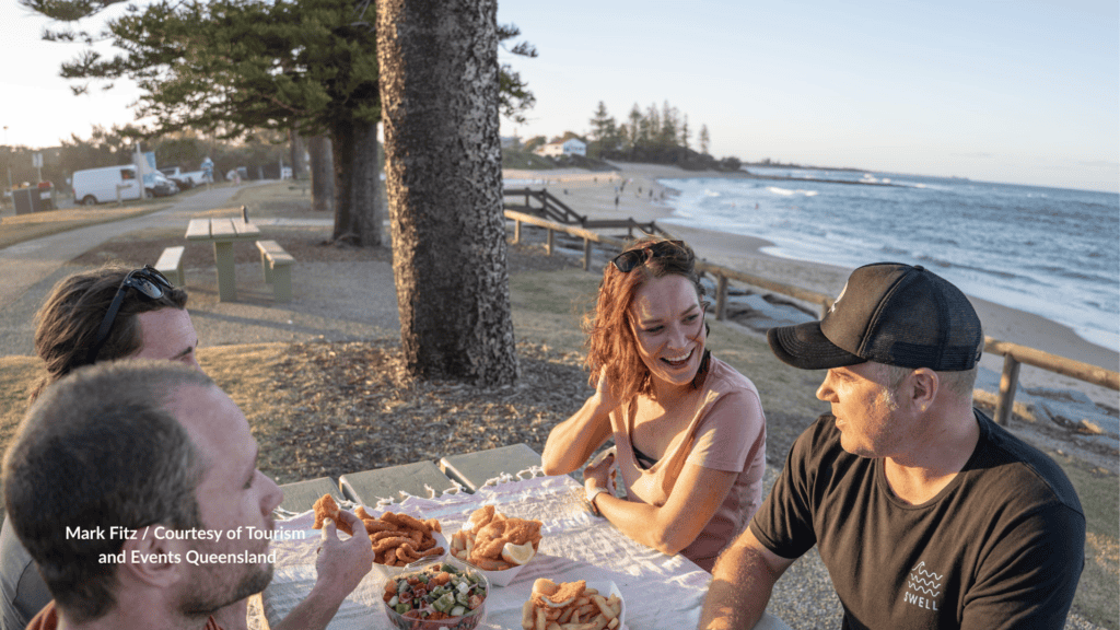Friends picnic at Moffat Beach Caloundra Queensland with fish and chips at a foreshore table looking toward the sand and Norfolk pines