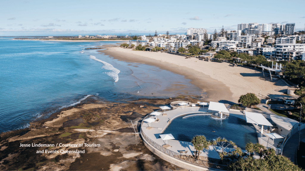 Aerial view of Kings Beach Caloundra Queensland with the beachfront pool in the foreground and the curved surf beach stretching past the apartments and Norfolk pines