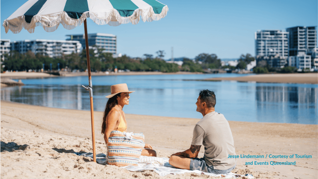 Couple sitting under a beach umbrella on the sand at Cotton Tree Beach Maroochydore Queensland looking across the calm Maroochy River