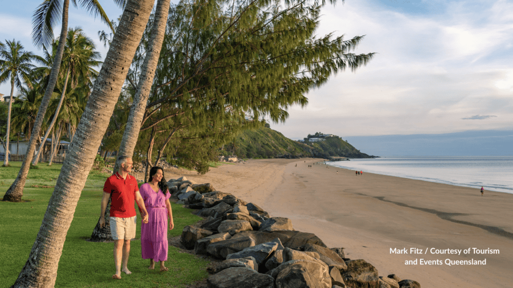 Couple walking under palm trees along the rock wall at Blacks Beach Mackay Queensland with the long shoreline and headland ahead