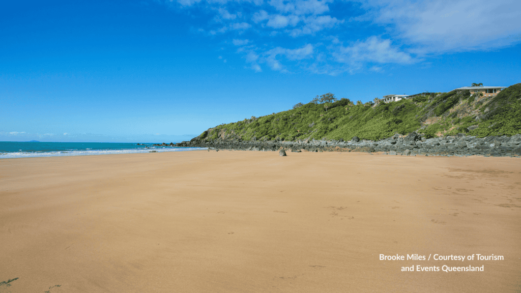 Wide golden sand at Sarina Beach Mackay Queensland with the rocky headland and houses on the green hillside under a clear blue sky