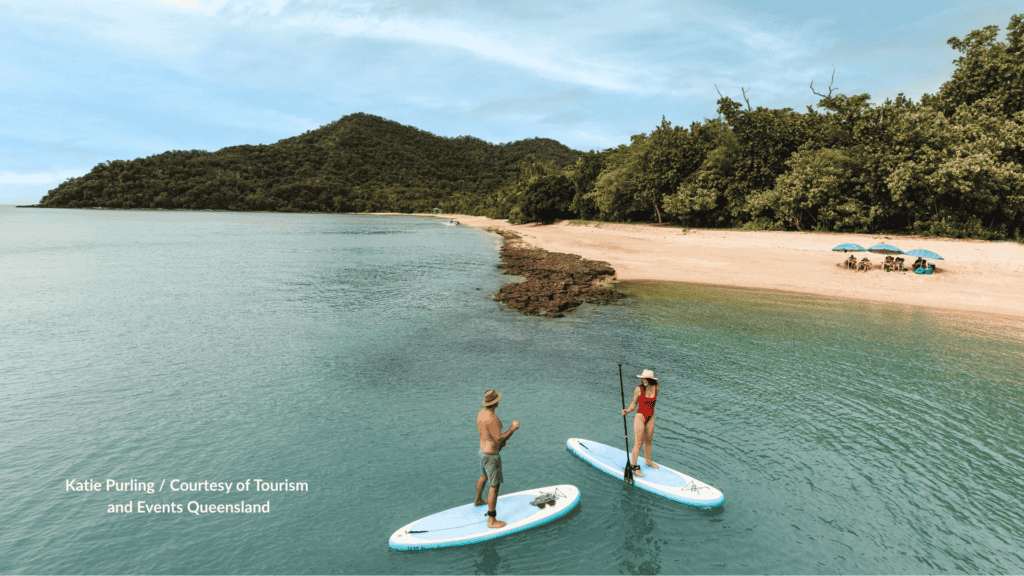 Man and woman stand up paddleboarding in calm water off Dunk Island near Mission Beach Queensland with rainforest hills and a sandy beach