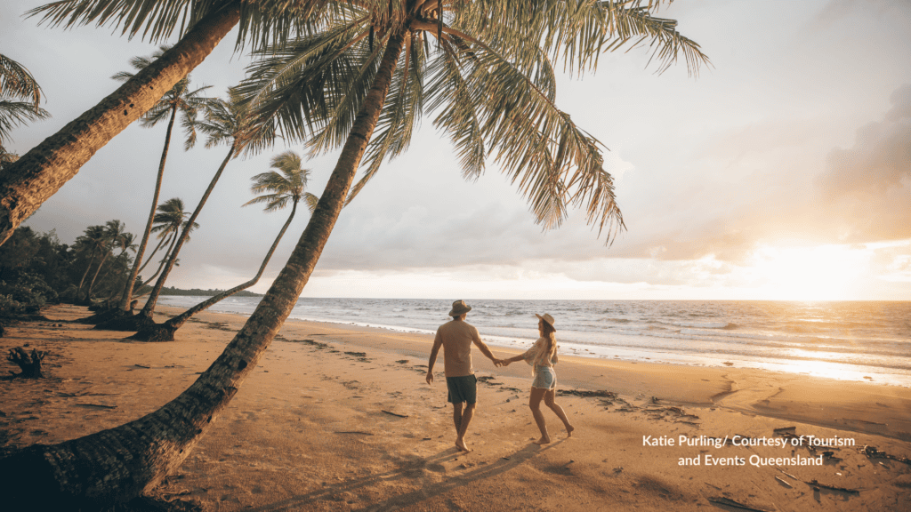 Couple walking hand in hand under leaning palm trees along Mission Beach Queensland at sunset