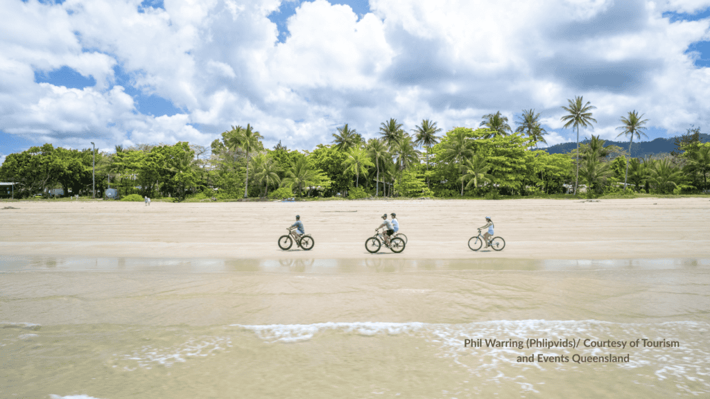 People riding bikes along the sand at Mission Beach Queensland with palm trees and rainforest behind