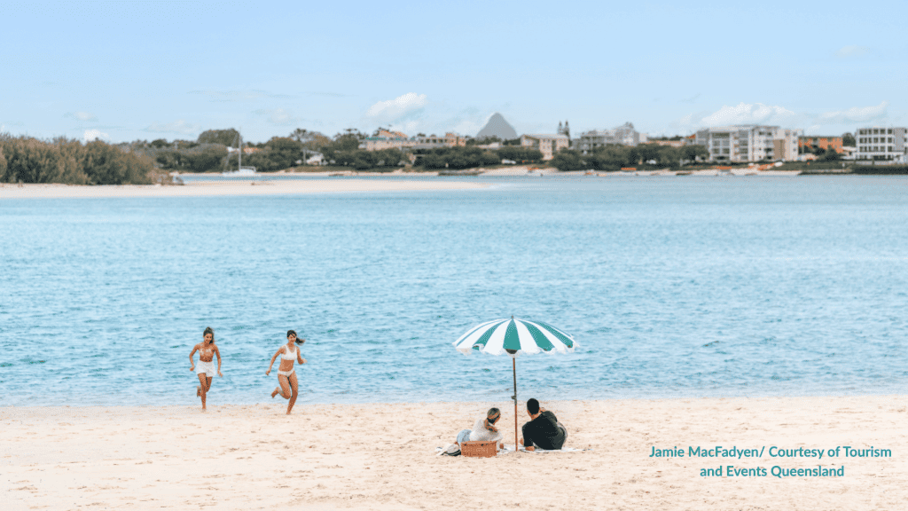 Kids running on Bulcock Beach Caloundra Queensland with a family under an umbrella on the calm passage sand