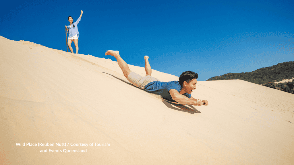 Man sand tobogganing down a dune at the Tangalooma Desert on Moreton Island Queensland while another guest stands at the top.