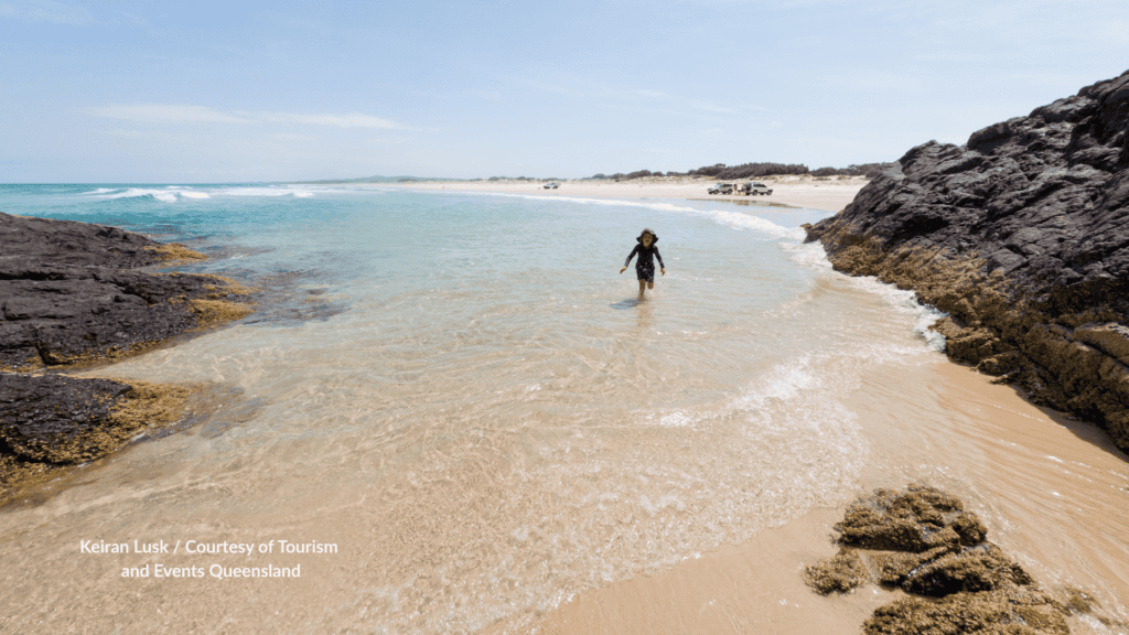 Child walking through a rocky tidal inlet at Eastern Surfside Beach on Moreton Island Queensland, with 4WDs parked on the ocean beach behind.