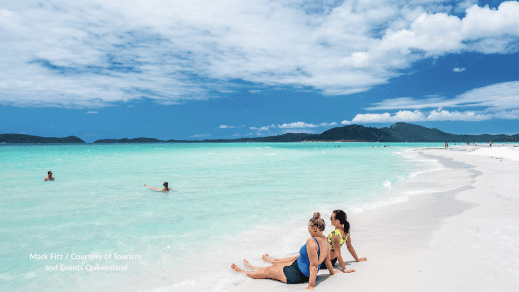 People sitting and swimming on the white silica sand and clear turquoise water at Whitehaven Beach on Whitsunday Island Queensland